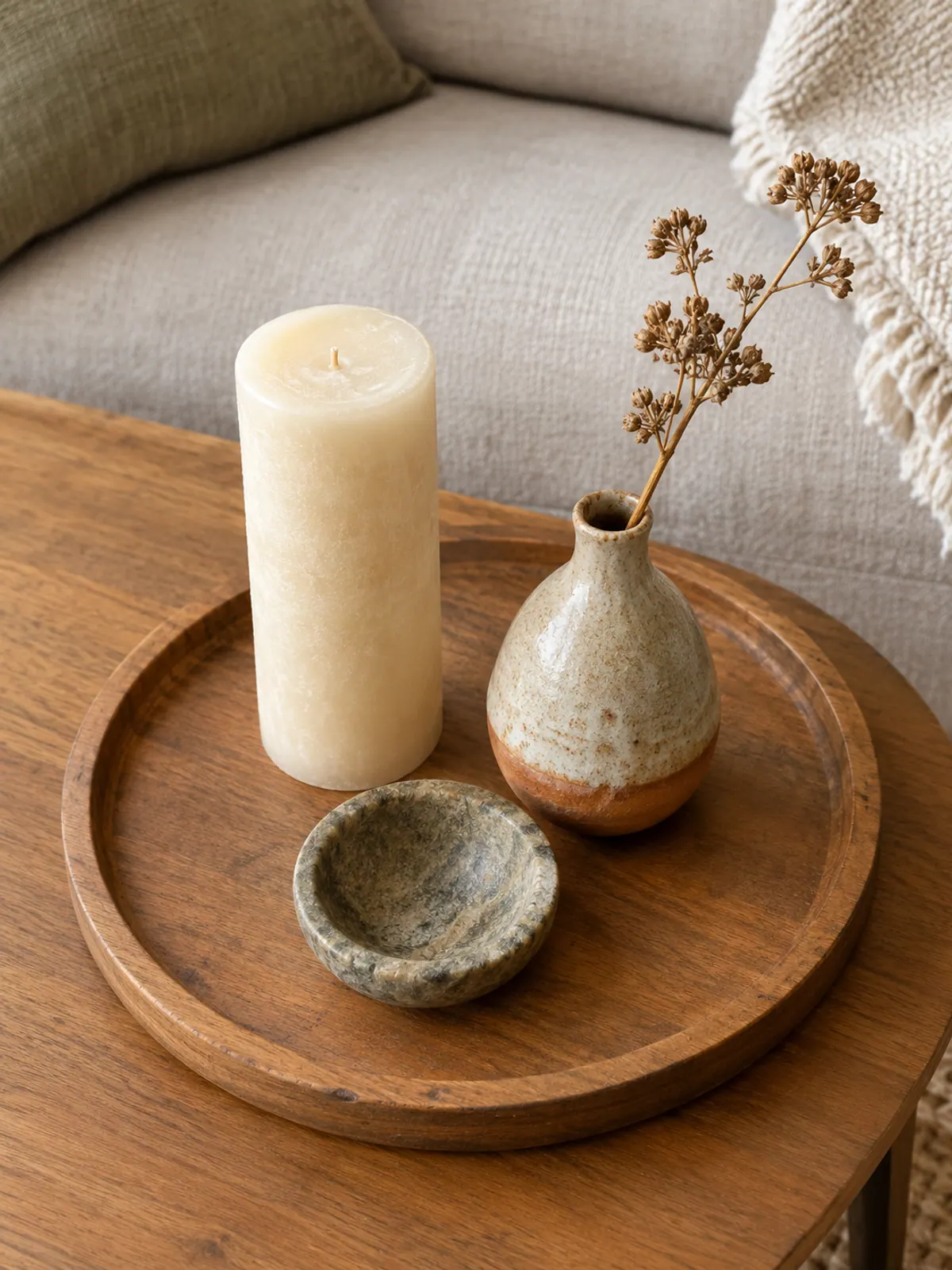 Round wooden tray holding a tall cream pillar candle, a ceramic bud vase with a dried stem and a small stone bowl